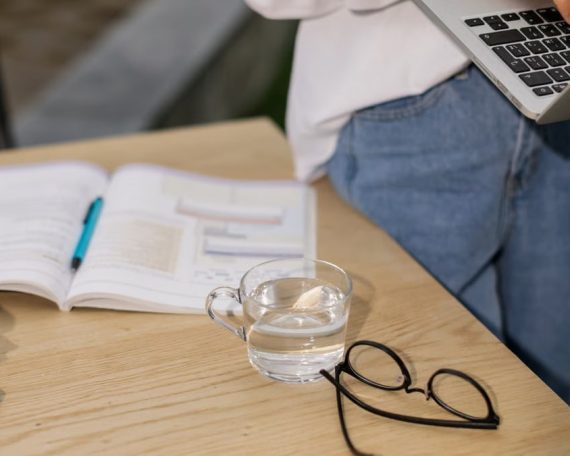 Casual indoor workspace with laptop, open book, eyeglasses, and a cup of water on a wooden table.